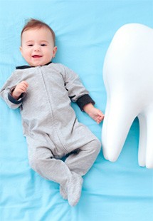 Baby in gray onesie next to giant plushy tooth on light blue sheet